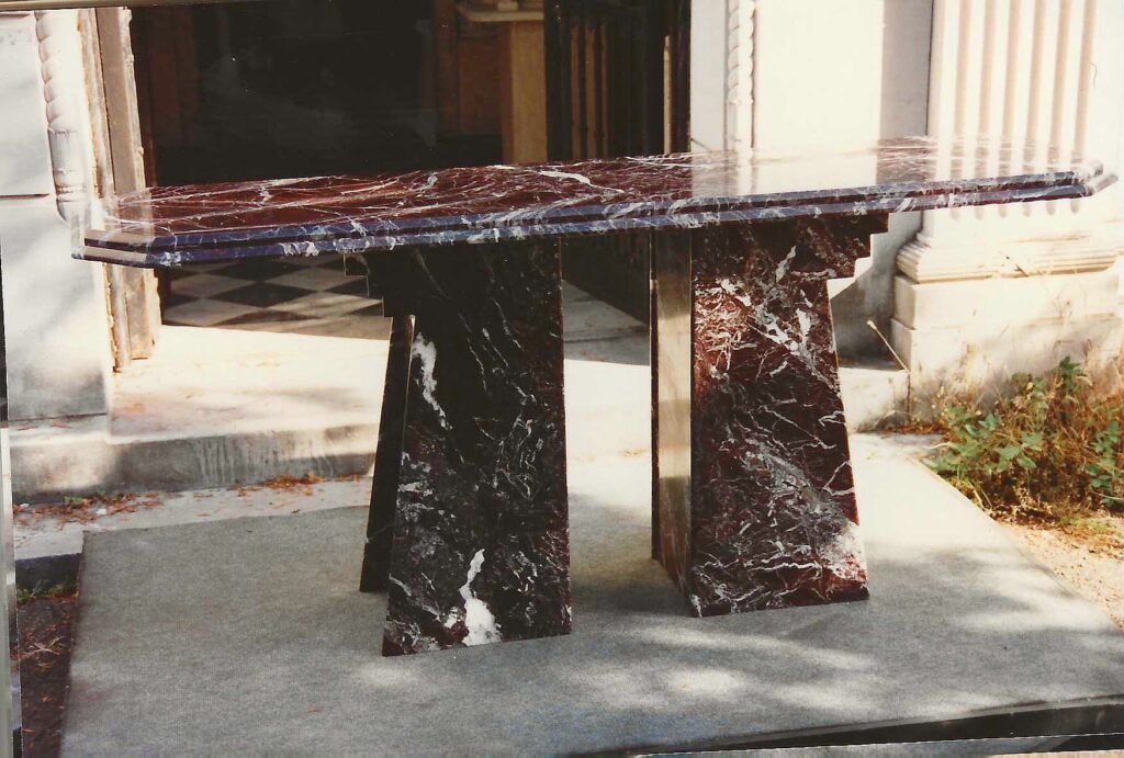 Bruce Bennett dining table in Rosso Levanto marble, photographed at Candoro Marble. The polished top is supported by paired tapered geometric pedestal bases.